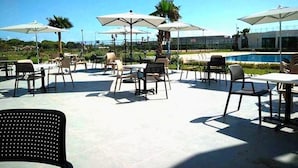 An outdoor patio area with tables and chairs, umbrellas, and a pool in the background. - Very nice apartment in Savannah Beach 4210 (Sidi Rahal)
