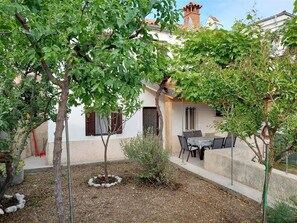 Une cour avec une table et des chaises, entourée d’arbres et bordée d’un bâtiment dont le toit est recouvert de tuiles.
