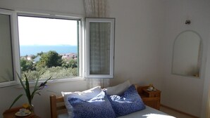 Ein Schlafzimmer mit Bett, Nachttisch, Spiegel und Fenster mit Blick auf das Meer und Gebäude.