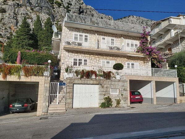A multi-story building with balconies, a stone facade, and a parking area with two cars.