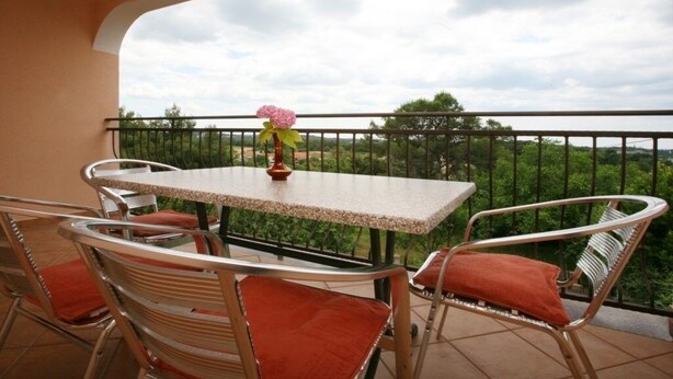 Un balcón con una mesa y sillas, un florero con flores, y vistas a zonas verdes y árboles.