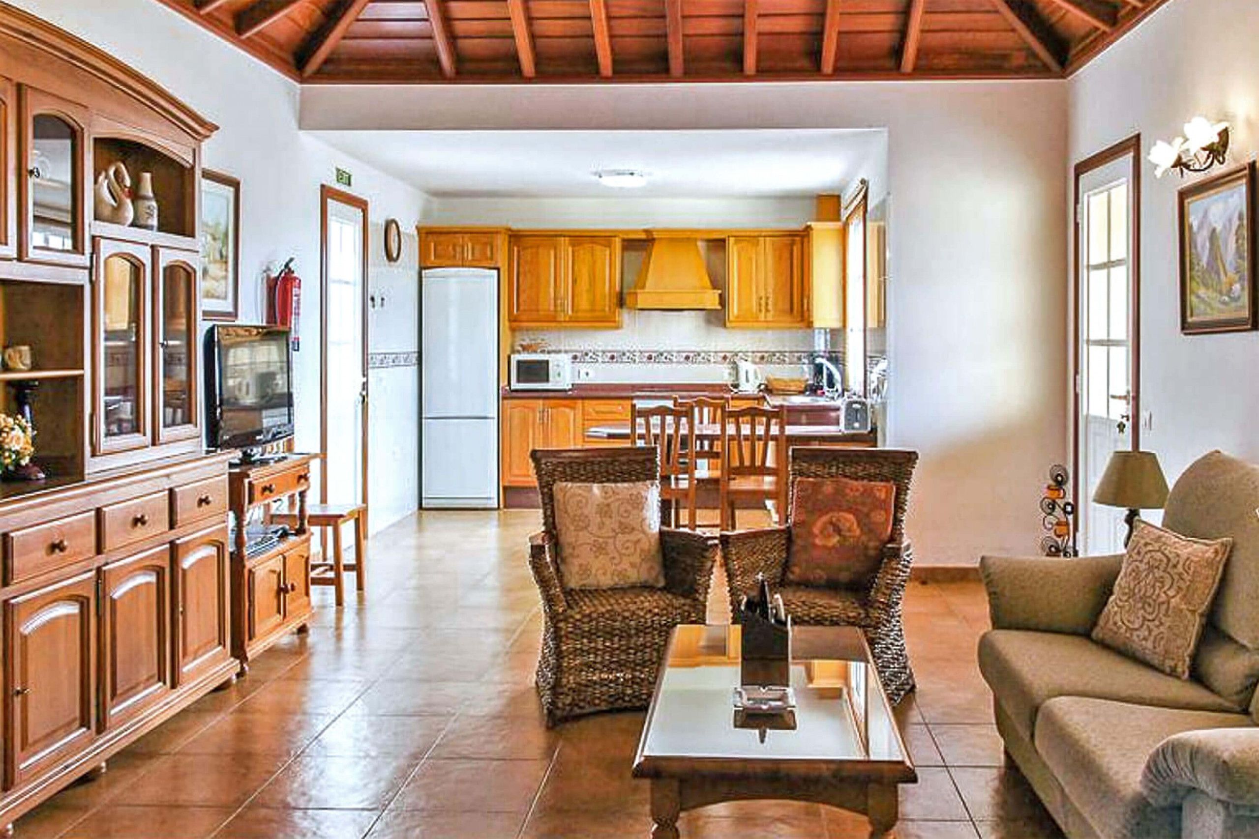 Amplia sala de estar con una mesa de comedor de madera, sillas de mimbre y un sofá. Se aprecia una cocina con armarios de madera y un refrigerador al fondo.