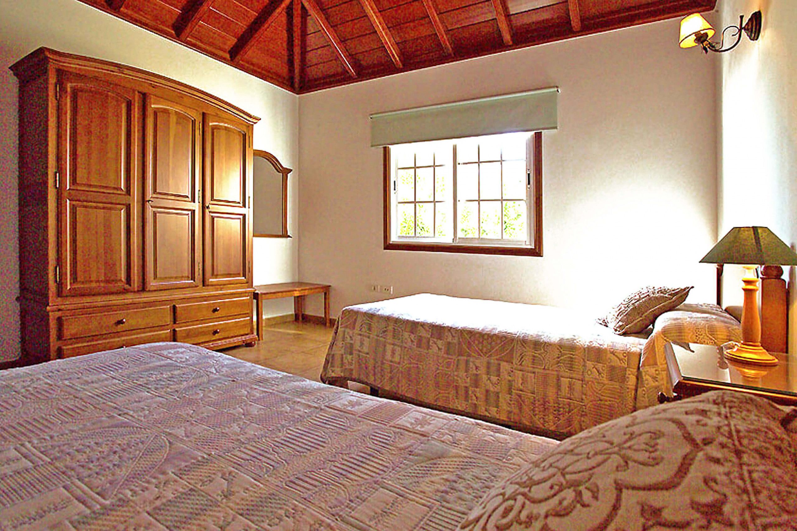 A bedroom with two beds, a wooden wardrobe, a window with blinds, and a lamp.