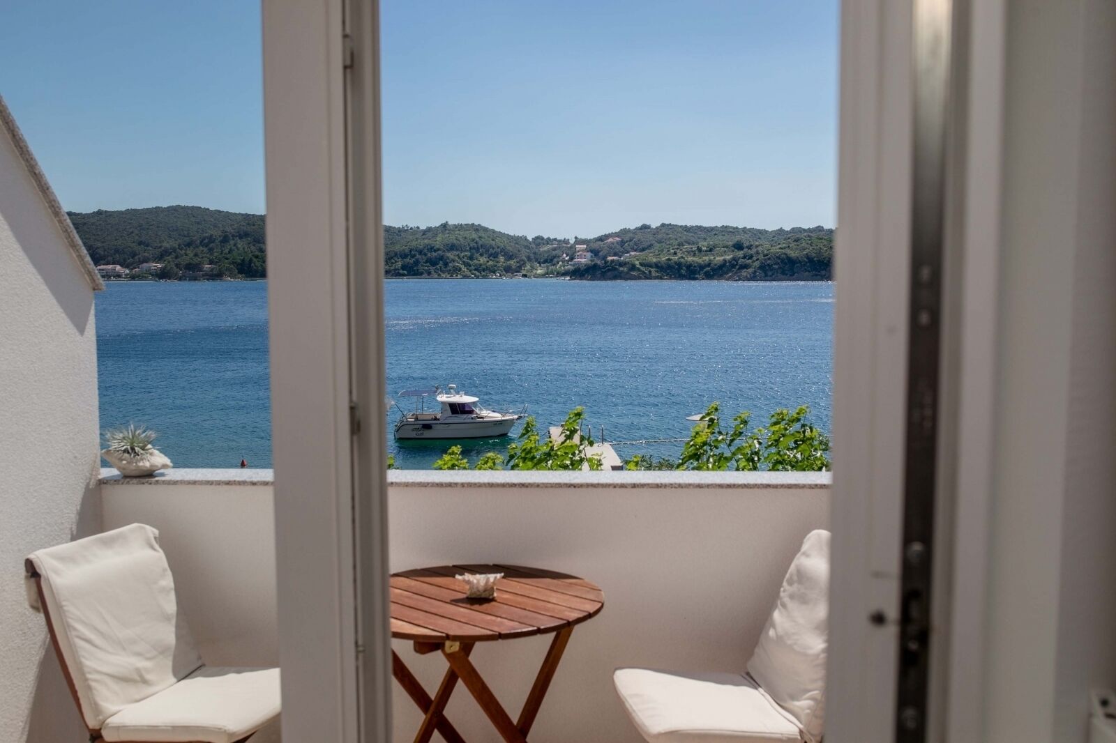 A balcony with a view of a lake and a boat, two white chairs, and a small wooden table.