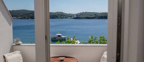 A balcony with a view of a lake and a boat, two white chairs, and a small wooden table.