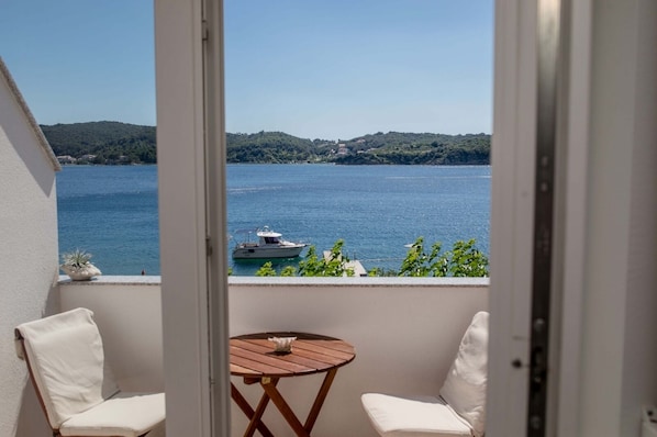 A balcony with a view of a lake and a boat, two white chairs, and a small wooden table.