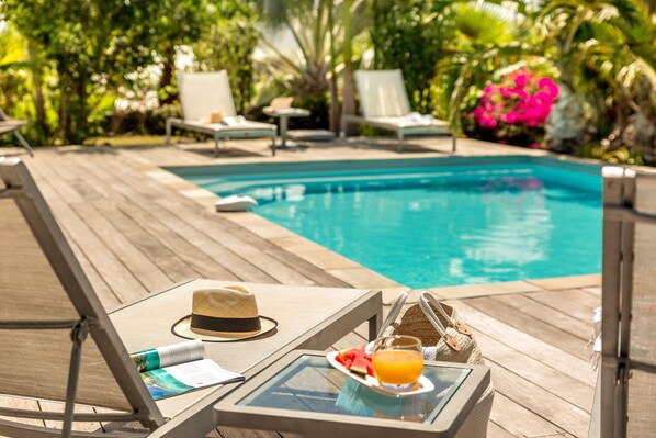 Un espace aménagé au bord de la piscine, avec des chaises longues, une table sur laquelle se trouve un chapeau, un verre de jus d’orange et un menu.