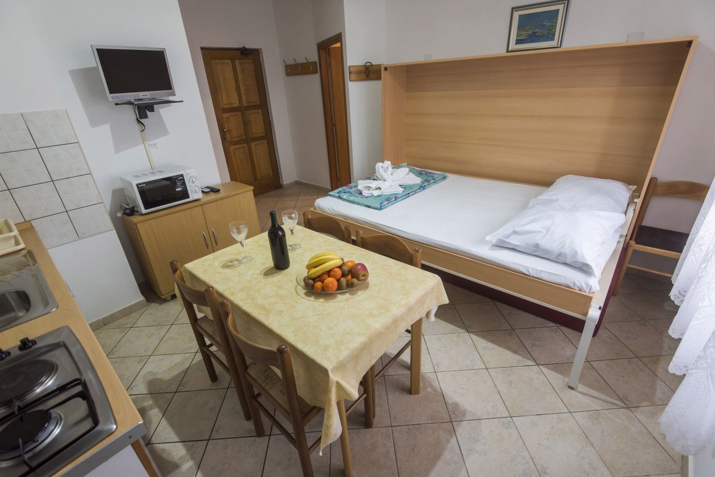A small, tidy room with a bed, a table with a fruit bowl, a microwave, and a television on the wall.