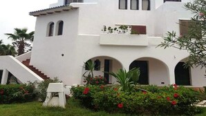 Un edificio blanco con una escalera que lleva a un nivel superior, rodeado de vegetación y flores rojas.