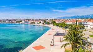 A coastal town with a clear blue sea, a promenade with palm trees, and buildings with red roofs. - Apartment in Novalja with Seaview, Terrace, Air condition, WIFI (3565-9) (Novalja)