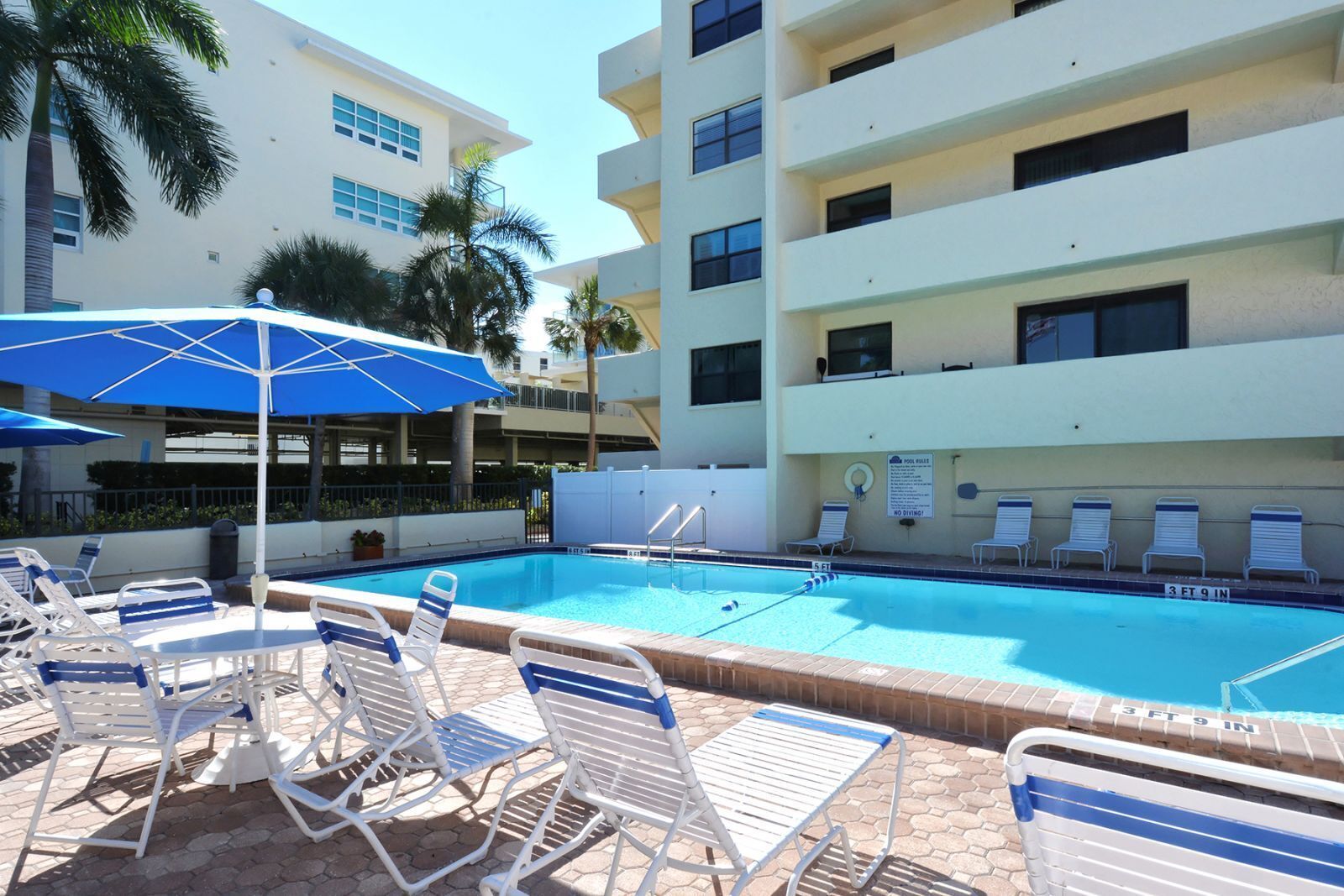 Une piscine entourée de chaises longues et de parasols, elle-même bordée d’un immeuble à plusieurs étages doté de balcons.