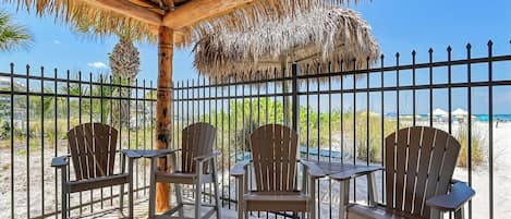 A covered outdoor seating area with chairs and tables, a thatched roof, and a view of the beach and palm trees.