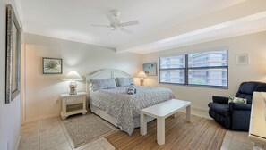 Une chambre à coucher comprenant un lit, des tables de chevet, un ventilateur de plafond, une fenêtre avec des stores et une chaise.