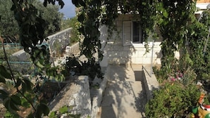 A stone pathway leading to a building with a window and shutters, surrounded by greenery and a stone wall.