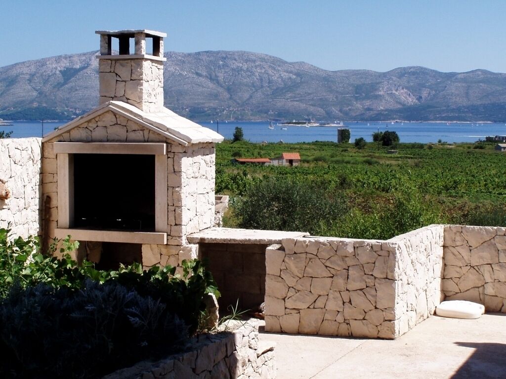 A stone-built outdoor kitchen with a chimney, surrounded by greenery and a view of a lake and mountains.