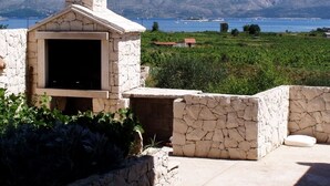 A stone-built outdoor kitchen with a chimney, surrounded by greenery and a view of a lake and mountains.