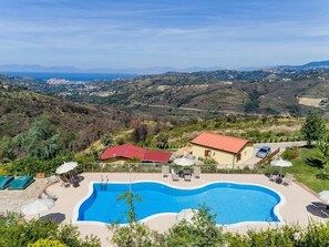 Ein Schwimmbad mit Liegestühlen und Sonnenschirmen, umgeben von Grün und mit Blick auf ferne Hügel und ein Gewässer.