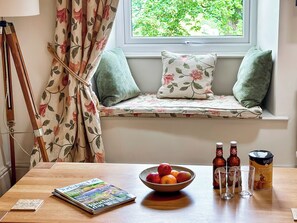 A cozy reading nook with a window seat, a wooden table with a bowl of fruit, a magazine, and a bottle of beer.