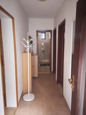 Ein schmaler Flur mit einer Holztür rechts, einer weißen Stehlampe und einem Badezimmer, das sich durch eine Glastür am Ende zeigt.