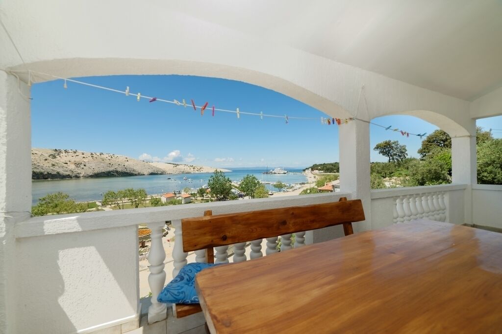 A balcony with a wooden table and bench, overlooking a coastal landscape with boats and hills.