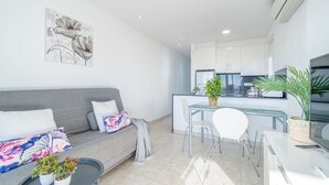 Un salon moderne avec un canapé gris, une petite table basse agrémentée de plantes, un coin repas avec une table et des chaises blanches, et une cuisine avec des meubles blancs et un four encastré.