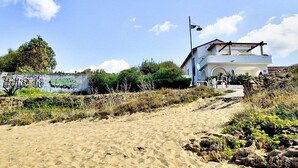 Un edificio blanco con techo inclinado y balcón, rodeado por una zona arenosa con vegetación escasa y un muro con grafitis.