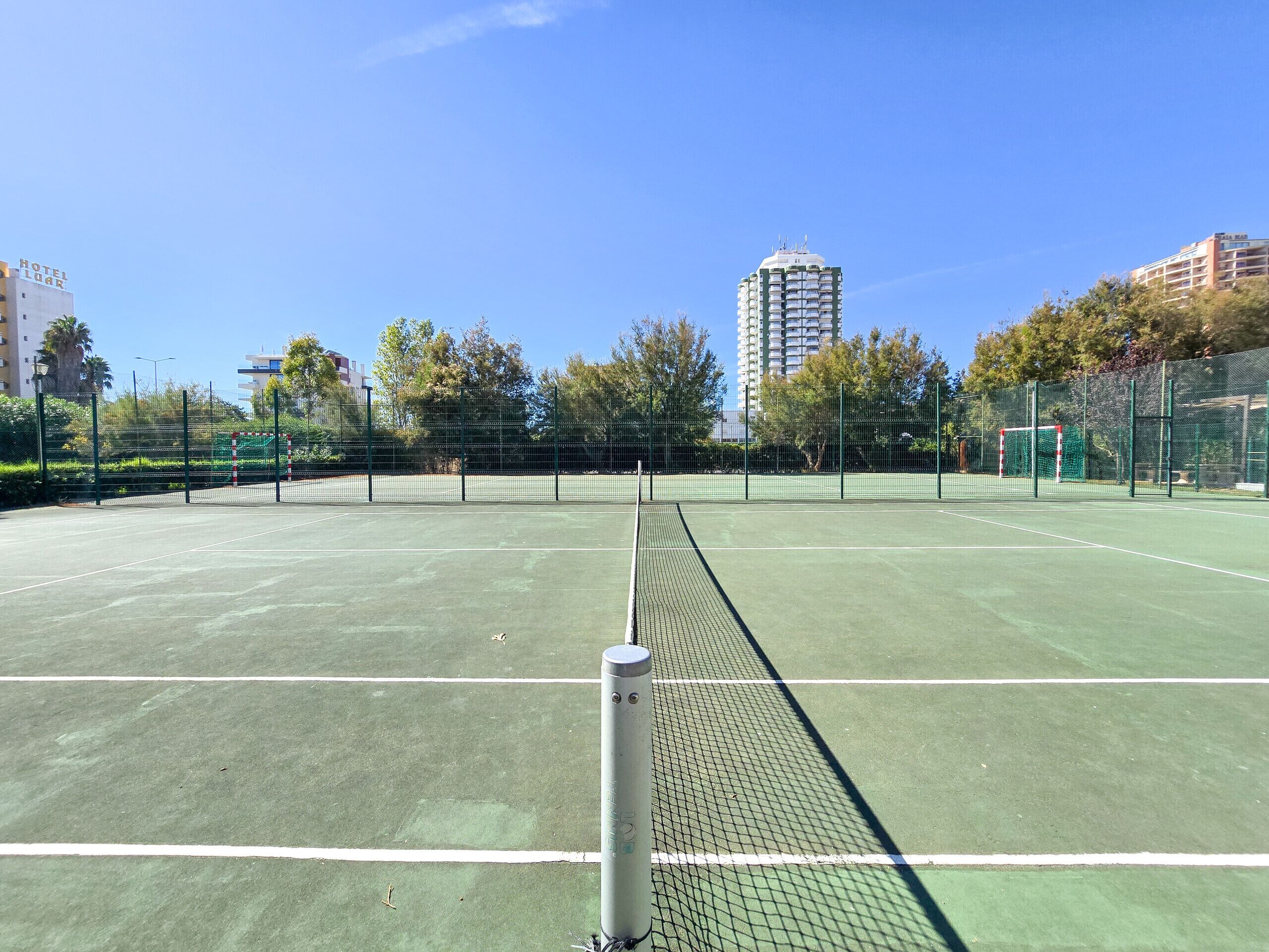 Una cancha de tenis con una red en el centro, rodeada por una valla y árboles.