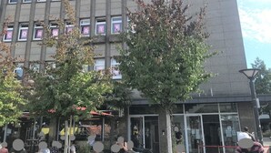 Il s’agit d’un bâtiment doté de nombreuses fenêtres et d’un toit plat. Des arbres se trouvent devant le bâtiment, et des personnes sont assises à des tables à l’extérieur.