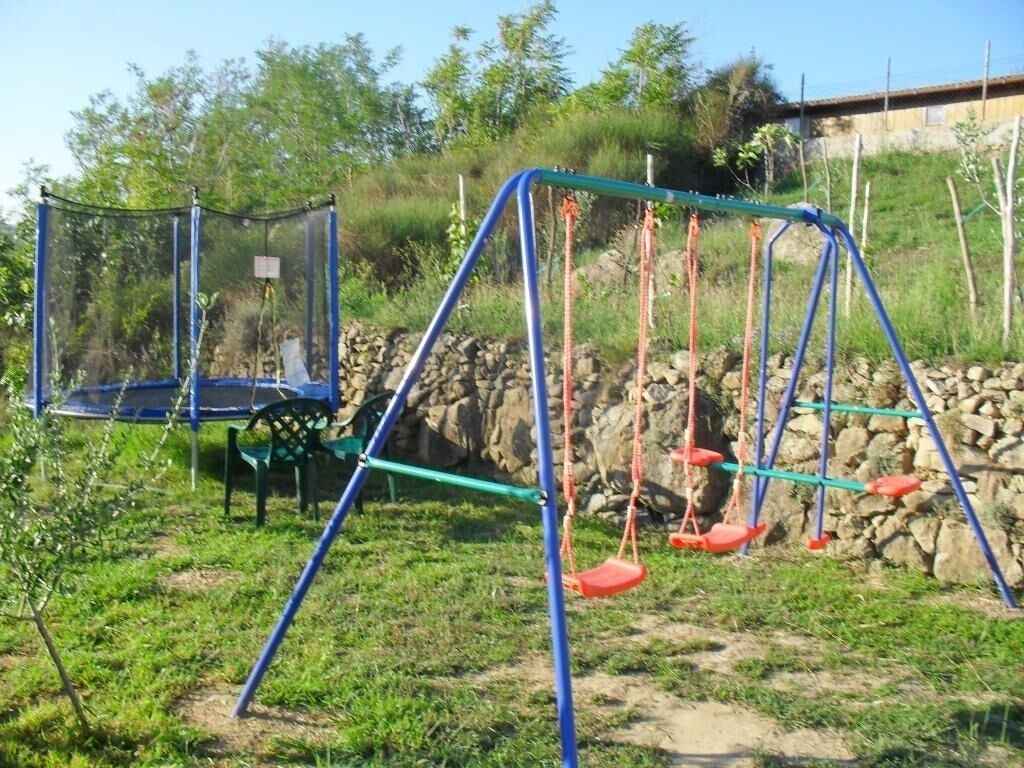 Un'area giochi con altalene e un trampolino, immersa nel verde e circondata da un muro in pietra.