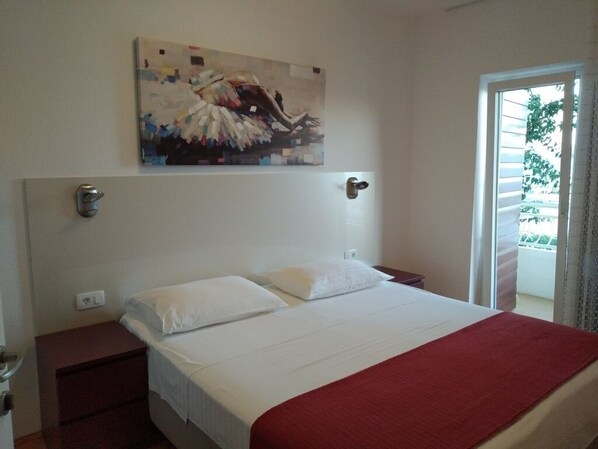 A bedroom with a bed, a nightstand, a painting on the wall, and a window with a view of greenery.