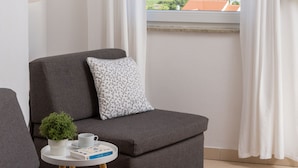 Ein modernes Zimmer mit grauem Sofa, einem kleinen Tisch mit Pflanze und Buch sowie einem Fenster mit Meerblick.