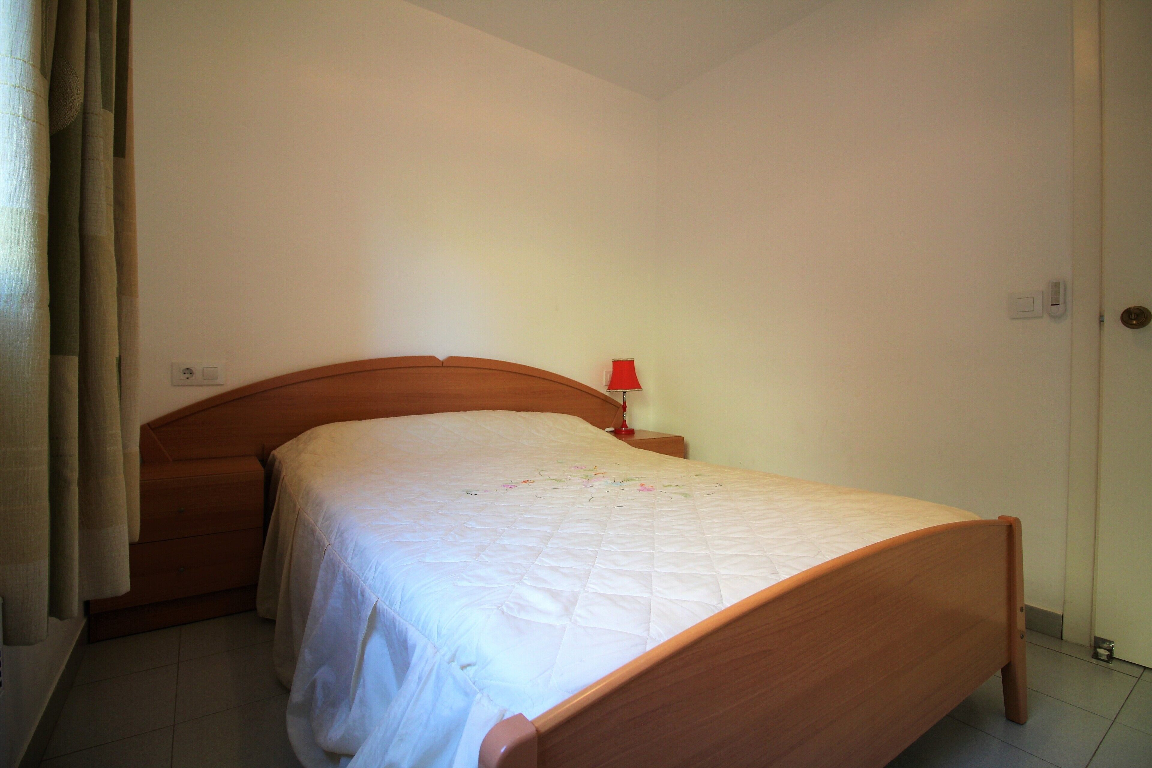 Uma cama de solteiro com cabeceira de madeira em quarto com paredes simples e chão de azulejos.