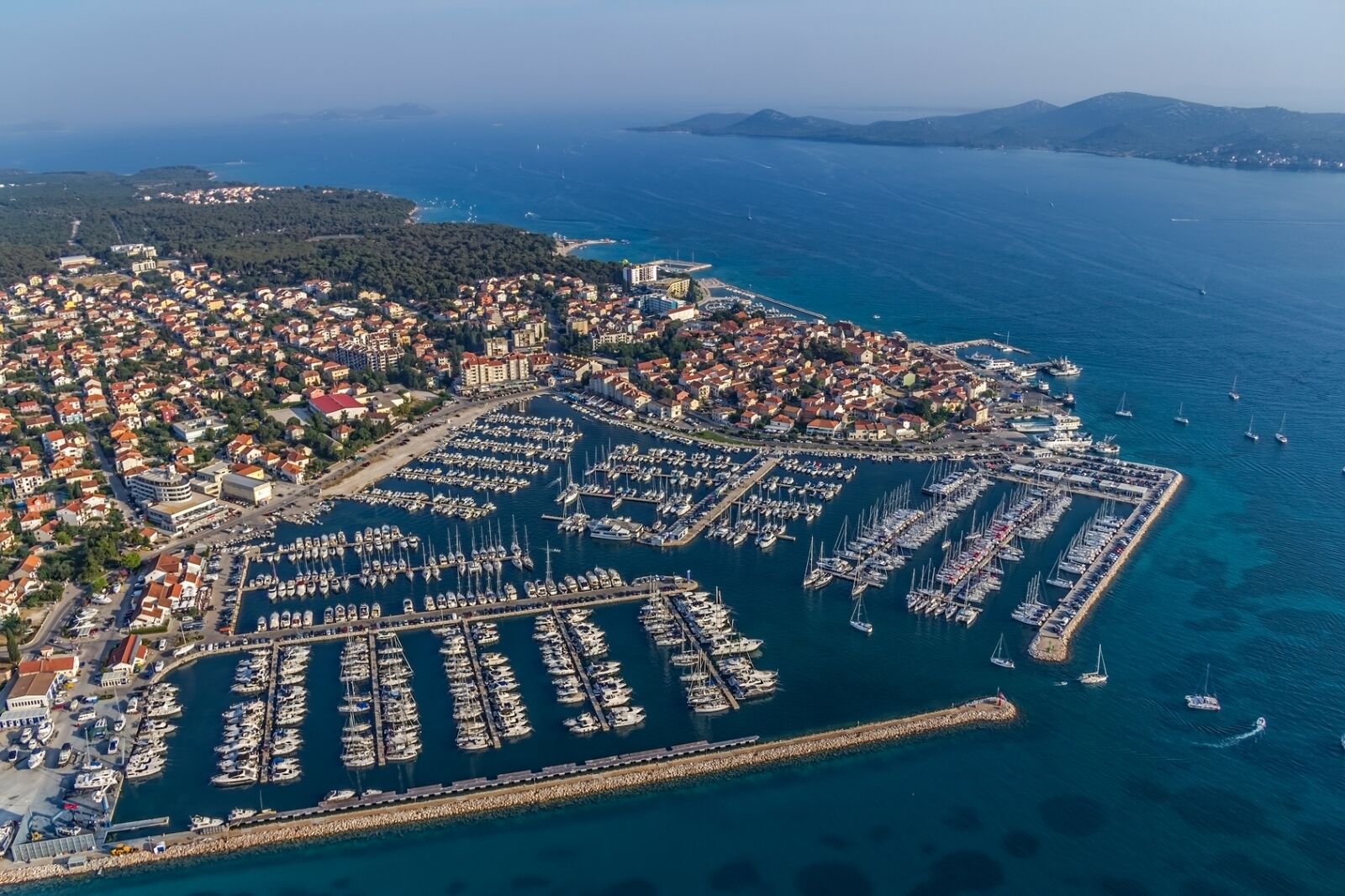 Une vue aérienne d’une ville côtière, avec un port de plaisance rempli de bateaux, entourée d’immeubles résidentiels et offrant une vue dégagée sur la mer.