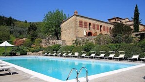 Ein Swimmingpool mit Liegestühlen, ein Gebäude aus Stein mit Bögen und eine Hügelkulisse.