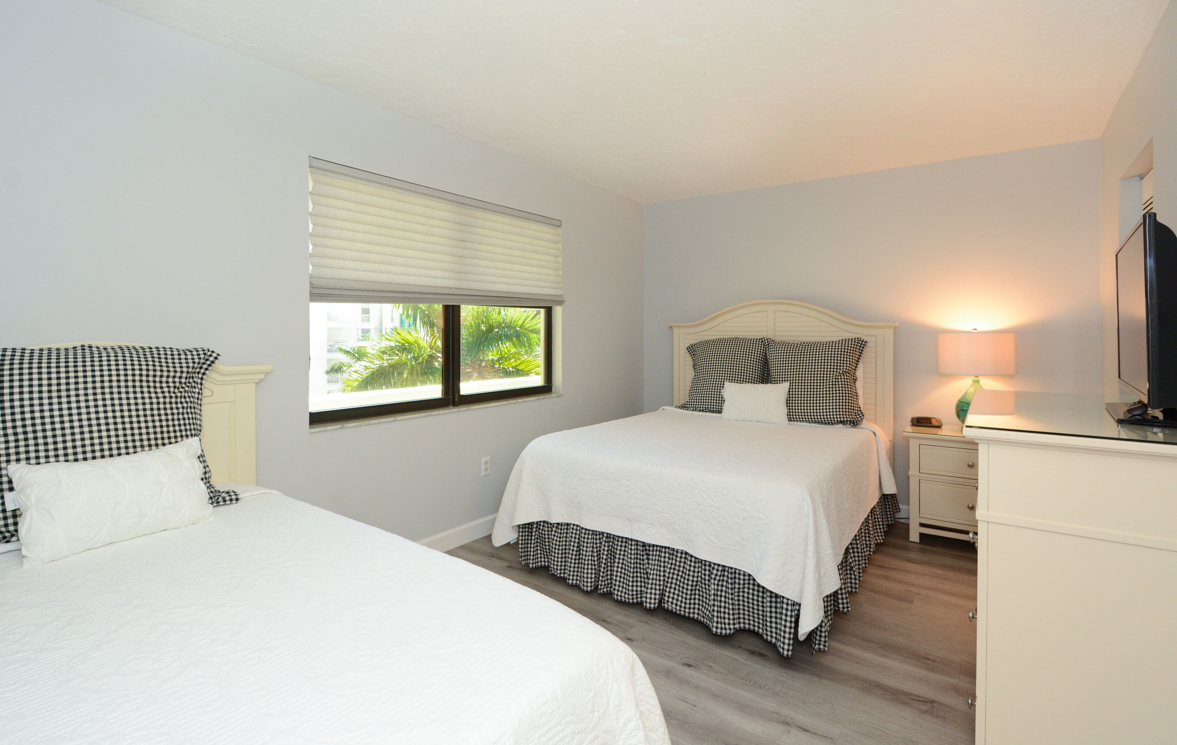 A bedroom with two beds, a nightstand, a dresser, a window with blinds, and a wall-mounted TV.