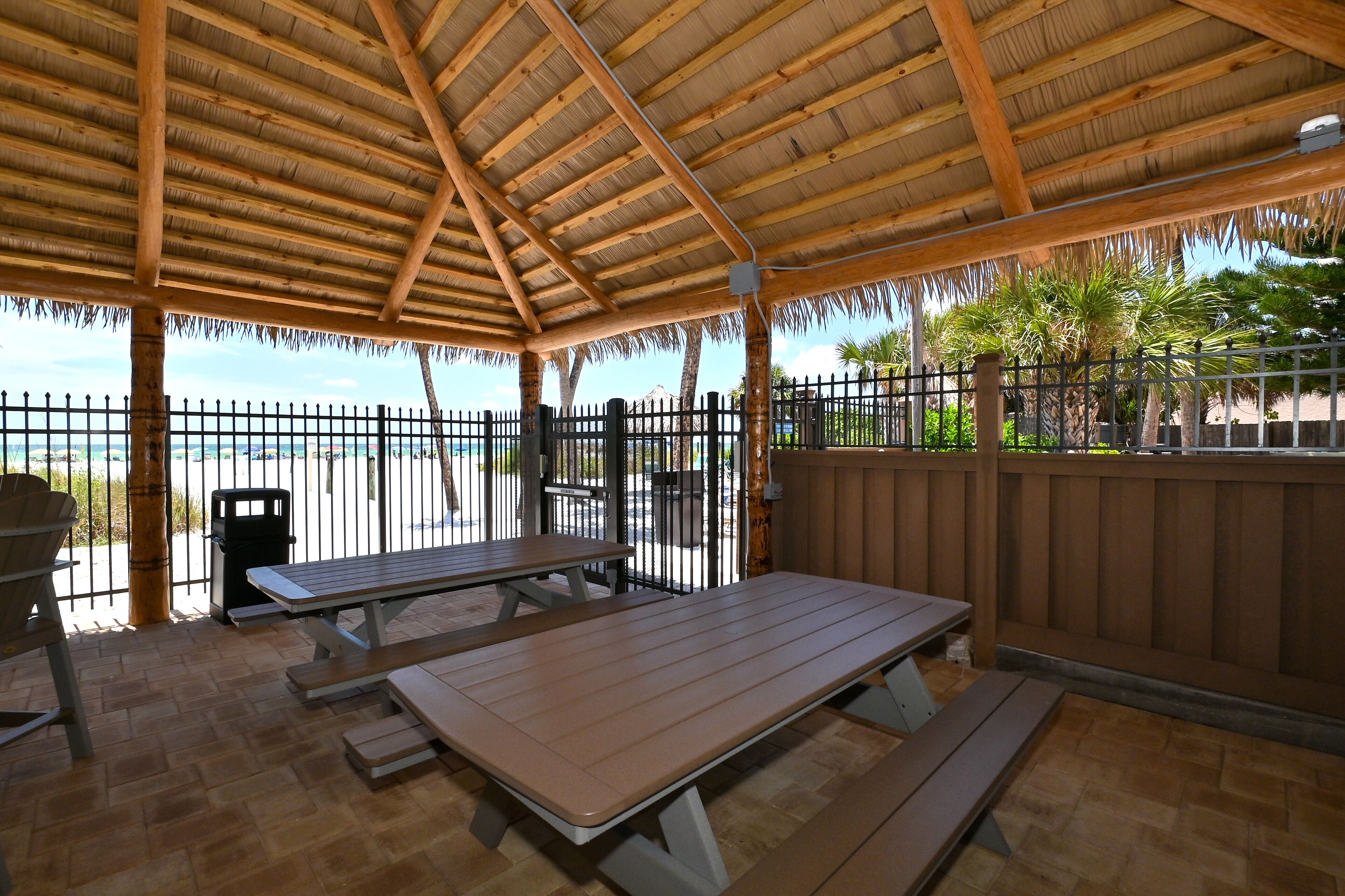 An outdoor patio with wooden tables and benches, a thatched roof, and a view of the sea.