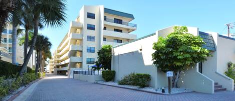 A paved walkway leading to a modern building with multiple floors and balconies, surrounded by palm trees and a clear sky.