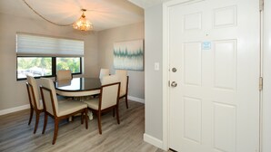 A dining area with a round table and four chairs, a chandelier, a window with blinds, and a door with a sign. - Steps to Siesta Key beach, restaurants and shops! (Sarasota)