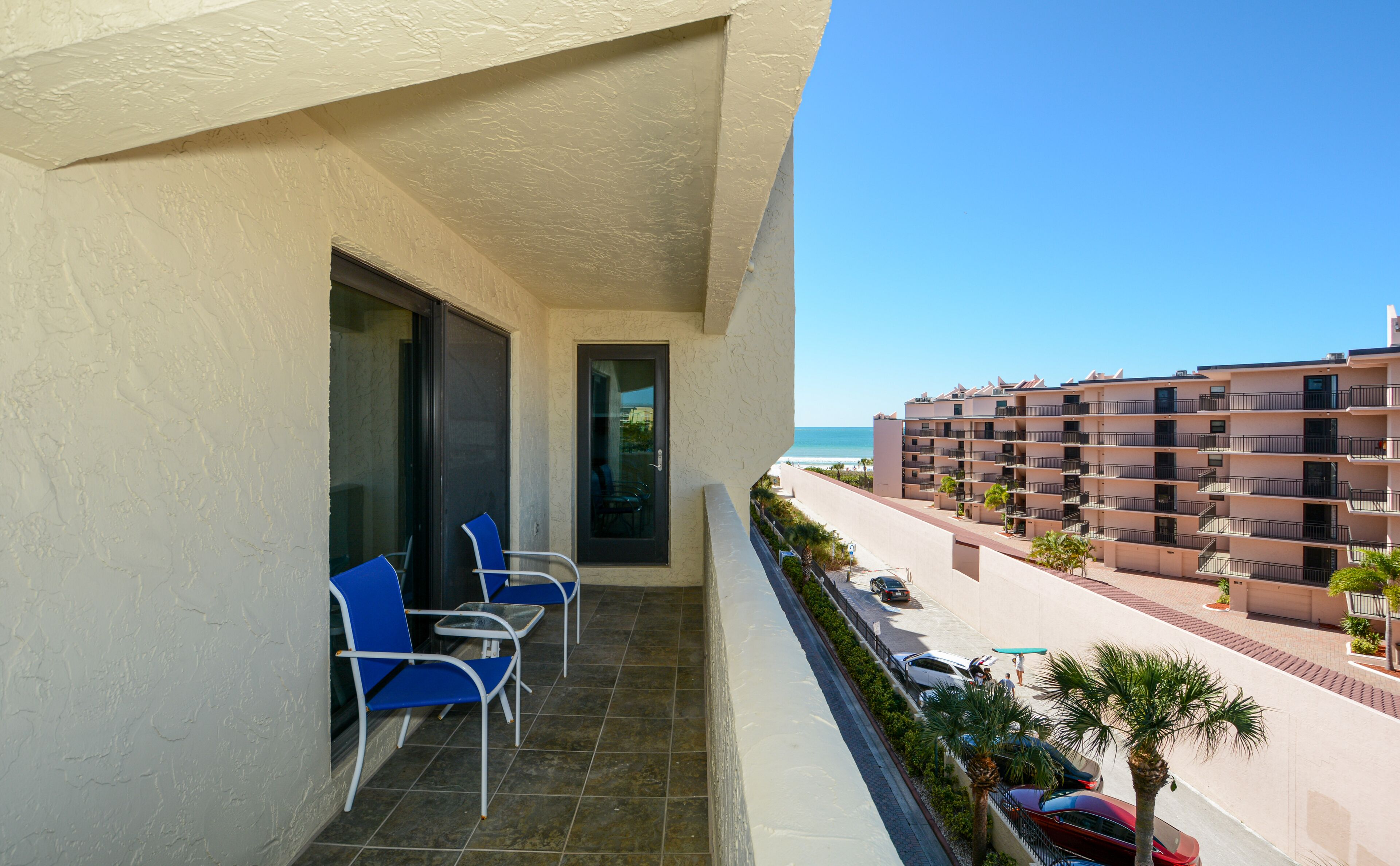 A balcony with blue chairs, a glass door, and a view of a beach and buildings.