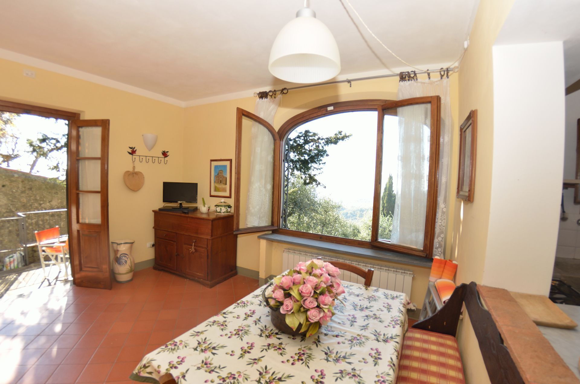 A bedroom with a bed, a dresser, a TV, a window with a view, and a table with a vase of flowers.