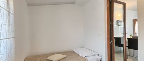 A small, single-bed room with a wooden door leading to a dining area.