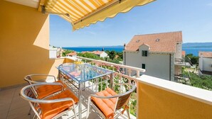 Ein Balkon mit einem Glastisch und orangefarbenen Stühlen, der auf eine Küstenstadt mit roten Ziegeldächern blickt.