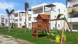 Une aire de jeux avec une structure de balançoire en bois et des balançoires jaunes, entourée d’une pelouse et d’un chemin pavé.