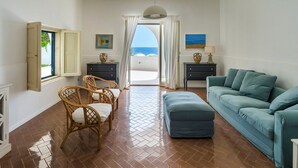 Un salon avec un canapé, des fauteuils, une petite table et une vue sur la mer, visible à travers une porte ouverte.
