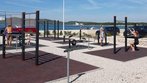 Un gimnasio al aire libre con diversos equipos de ejercicio, incluyendo barras de dominadas y máquinas de pesas, ubicado sobre una superficie de grava y con una vista despejada al mar.