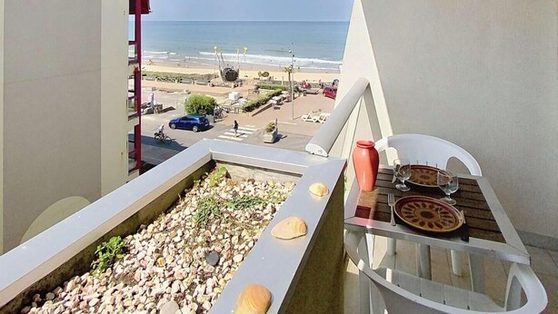 Un balcón con vista a la playa, una mesa de comedor con un florero y platos, y una pila de piedras.