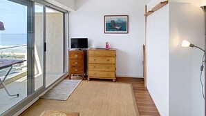 Habitación con un amplio ventanal corredero que da a un balcón con vista al mar. Hay un aparador de madera con cajones, una mesita con un posavasos tejido y una lámpara de pie.