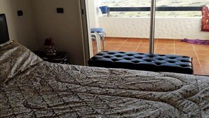 Un dormitorio con colcha estampada, un banco tapizado negro y vista a un balcón con barandilla blanca y un paño rojo.
