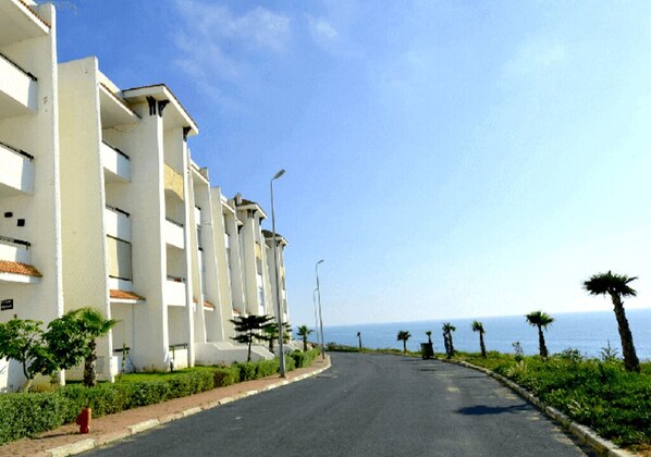 A coastal road lined with white buildings, palm trees, and a clear view of the sea.