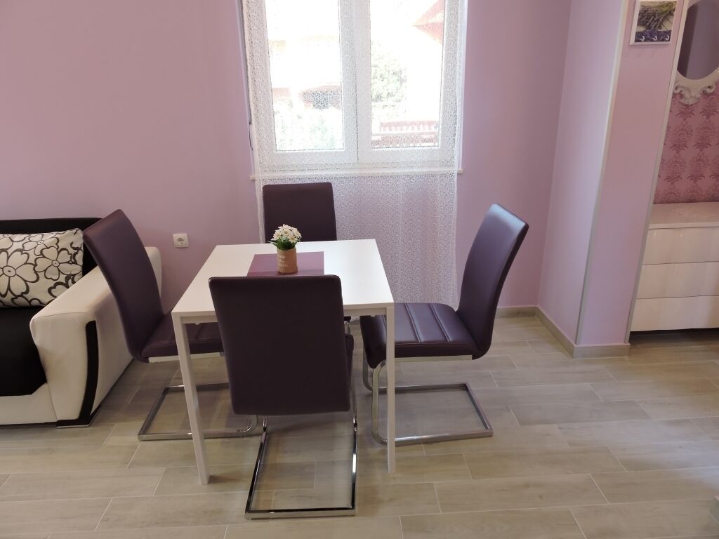 Un comedor con una mesa blanca y cuatro sillas, un jarrón pequeño con flores, y una ventana con vistas al exterior.